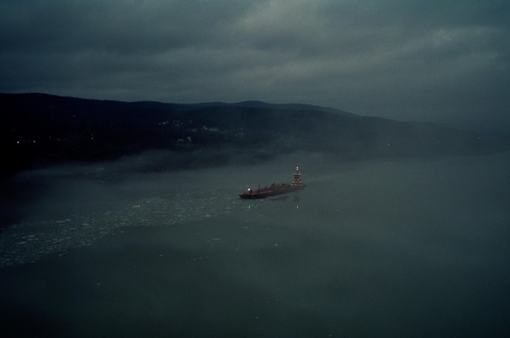 A ship with illuminated lights navigates through a foggy body of water with ice floes, with dark hills in the background.