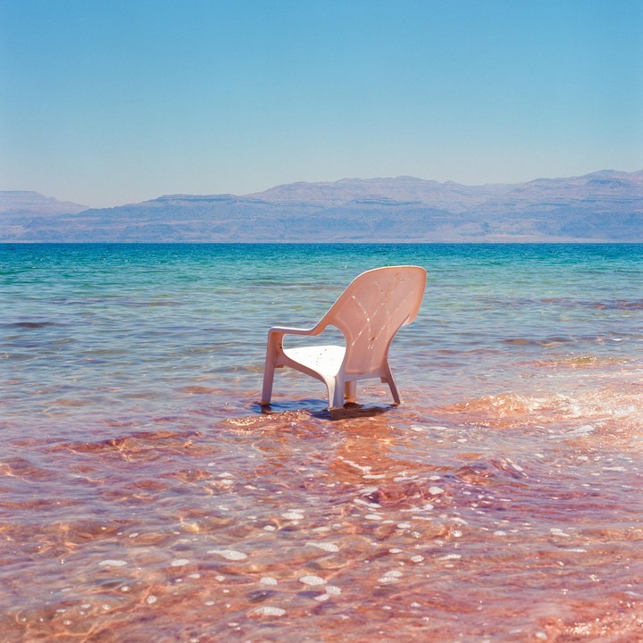 A white plastic chair sits in shallow, reddish-brown water with a blue sea and mountains in the background.