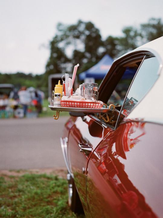 A red car with a tray of food and drinks hanging from its window is parked on a grassy area.