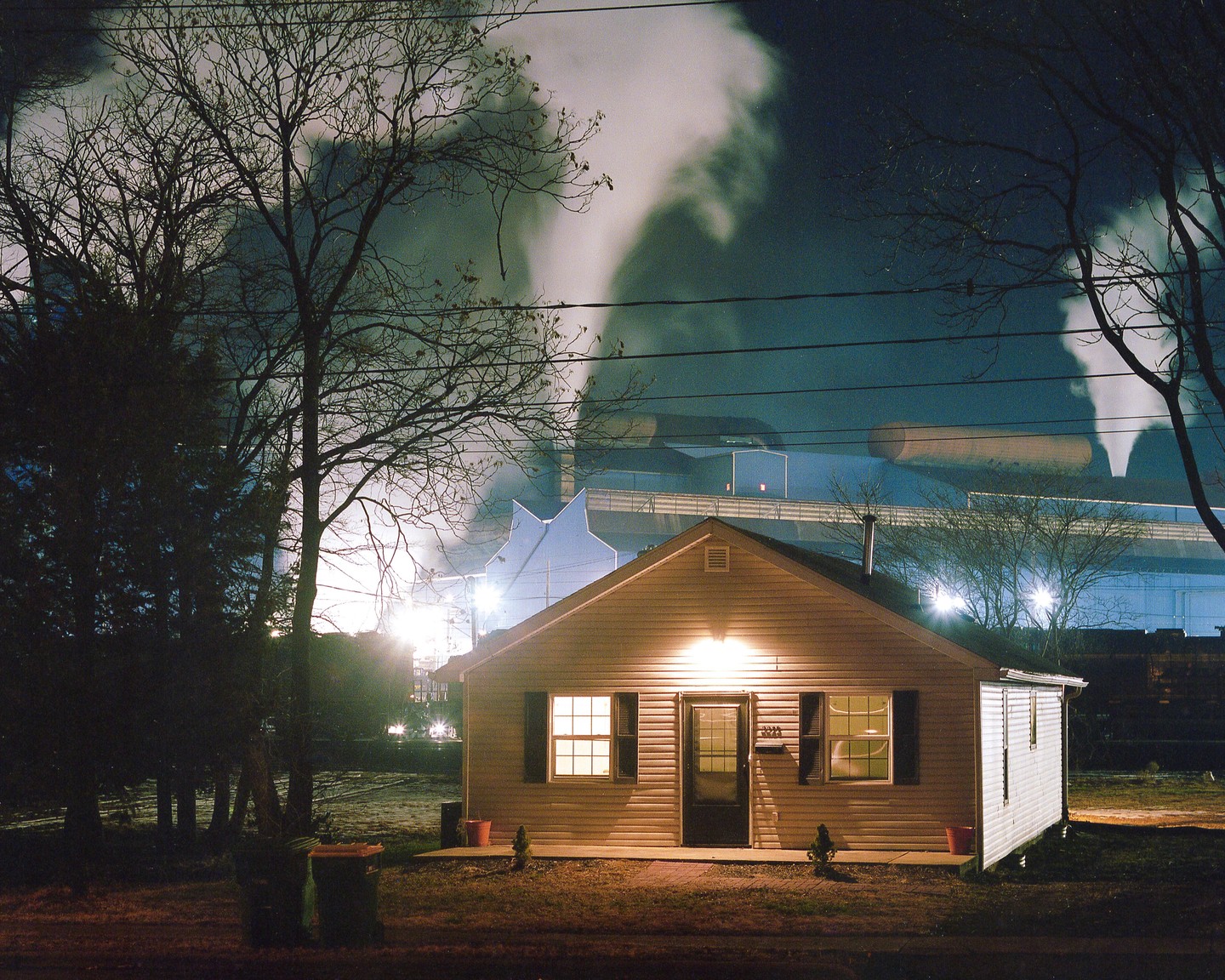 A small house with illuminated windows stands in front of a large industrial complex emitting smoke into the night sky.