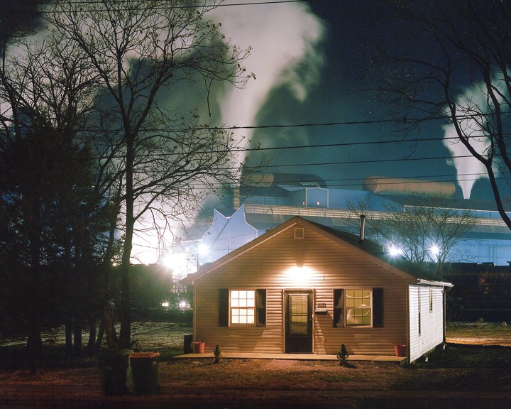 A small house with illuminated windows stands in front of a large industrial complex emitting smoke into the night sky.