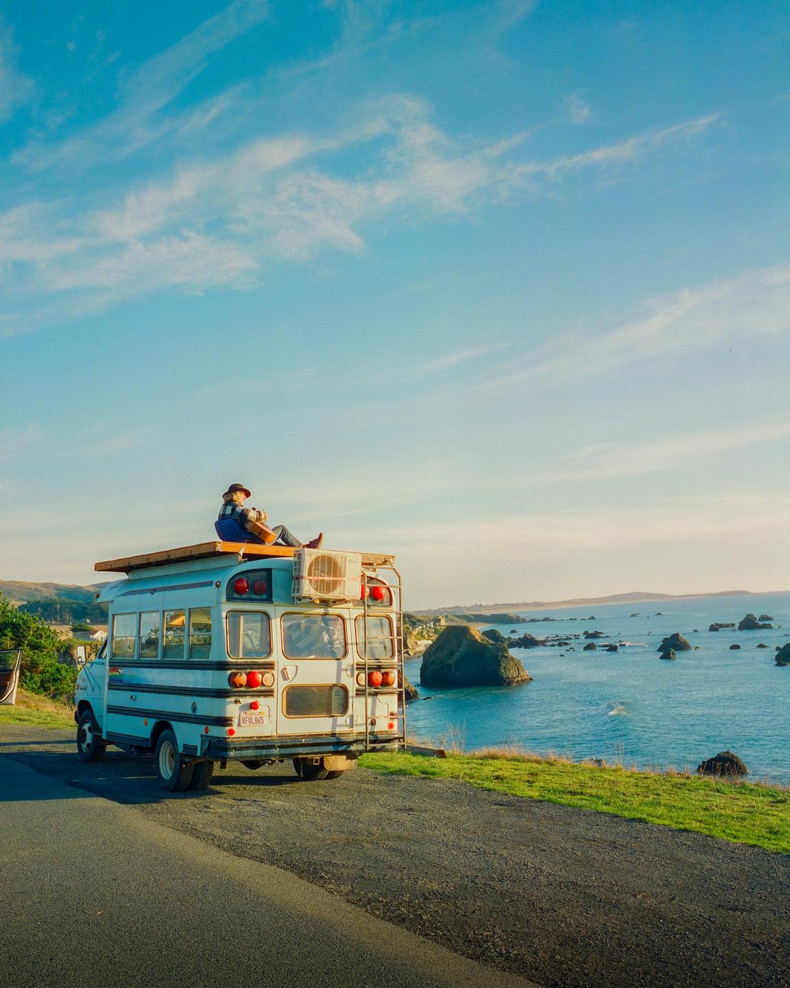A person sits on the roof of a converted bus parked on a road overlooking the ocean with rock formations.