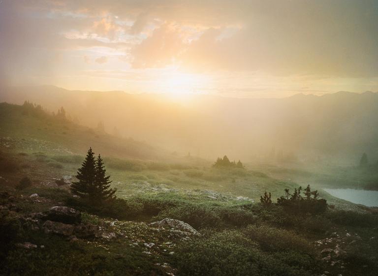 The sun sets over a hazy mountain landscape with a small lake and evergreen trees.