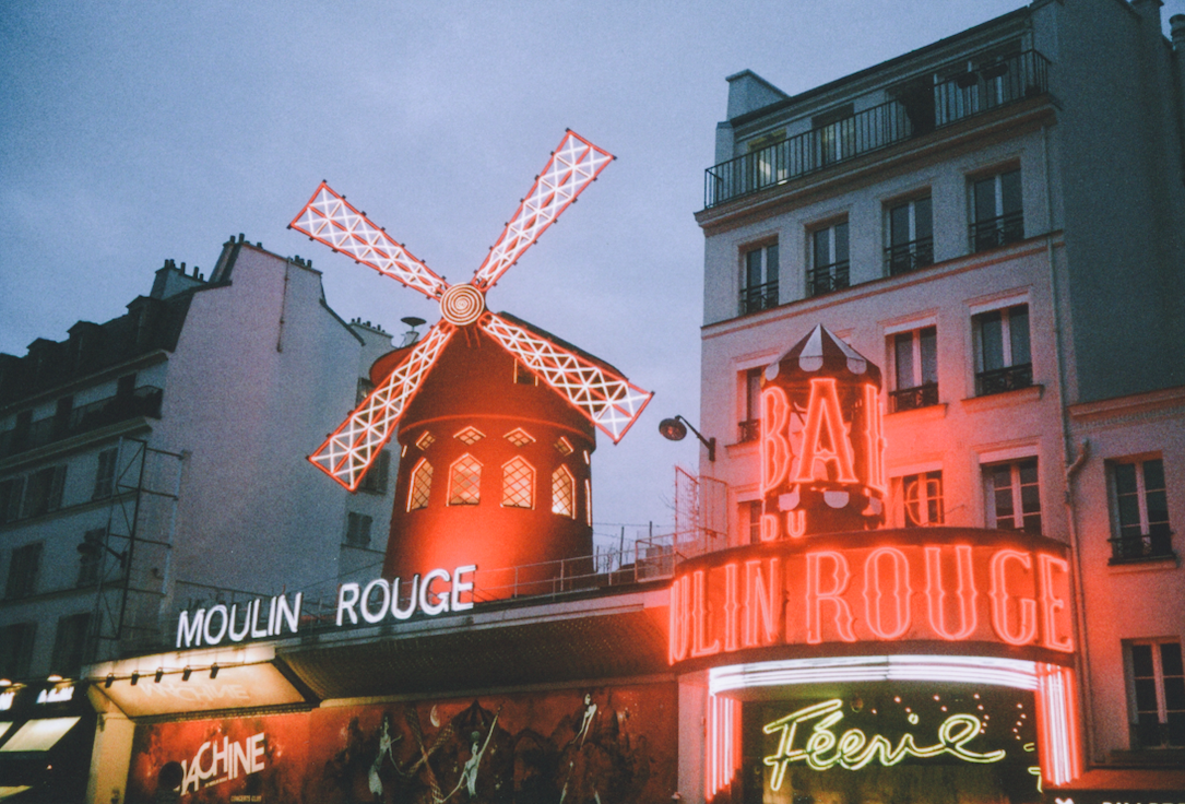 The Moulin Rouge building with its iconic windmill and neon signs is visible at dusk.