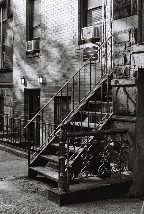 A black and white photo shows an exterior staircase with ornate railings leading up to a brick building with windows and air conditioning units.