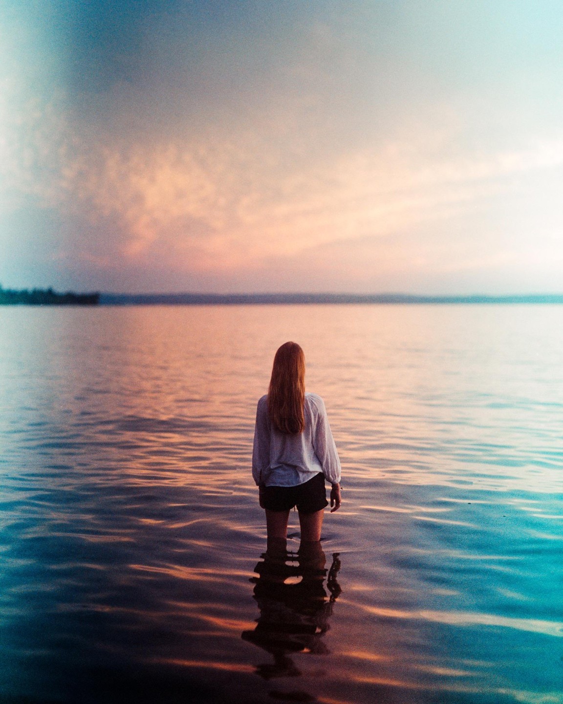 A woman stands in a body of water with a colorful sky at sunset.