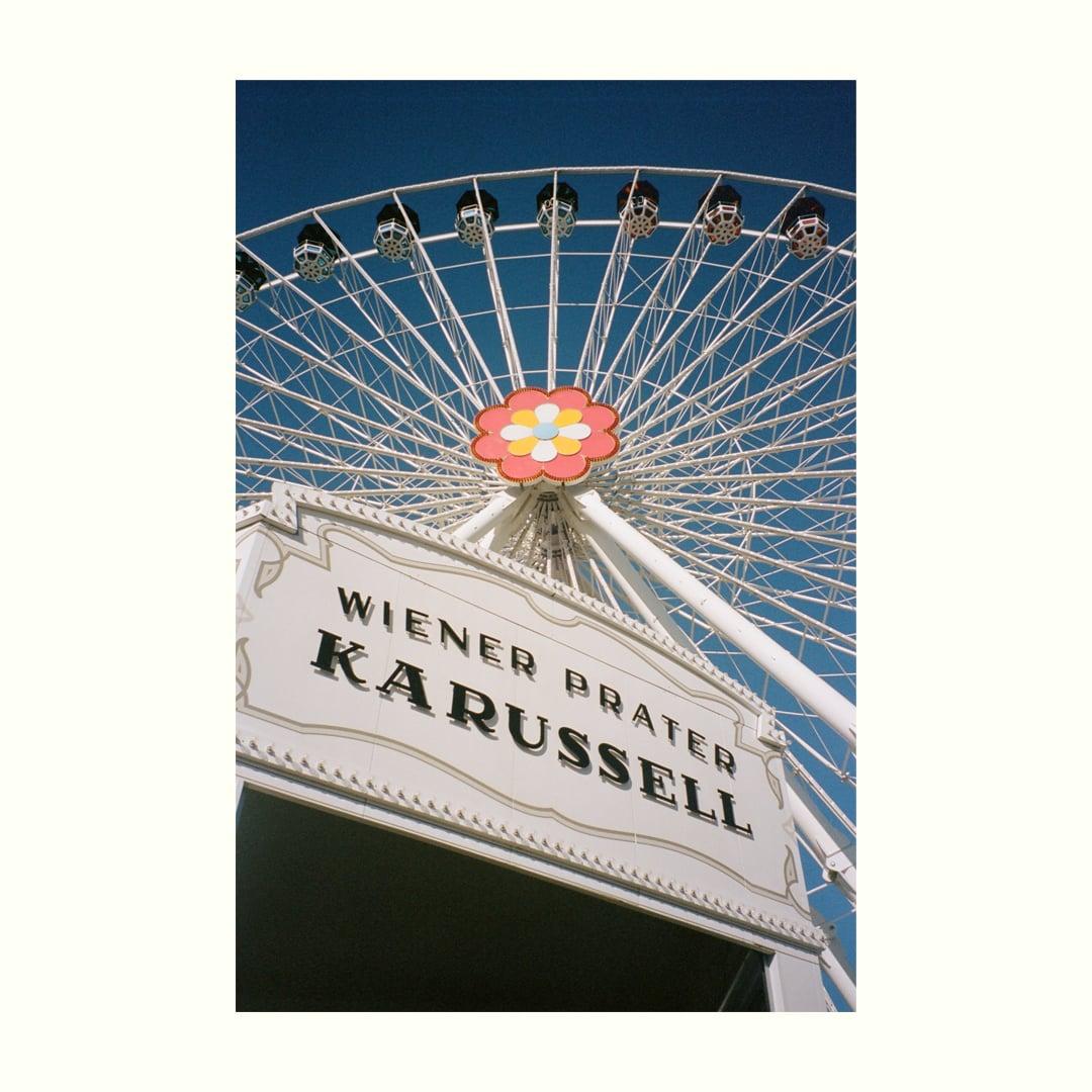 A low-angle view shows a white ferris wheel with a colorful flower design at its center, set against a clear blue sky.