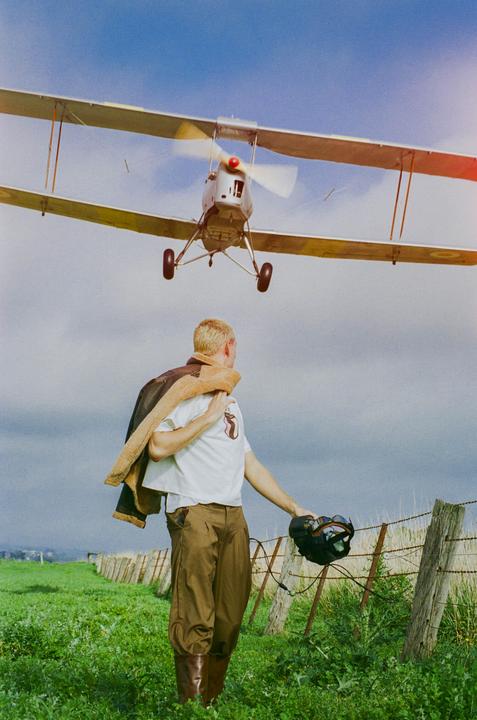 A man in a pilot jacket and boots stands in a field looking at a biplane flying overhead.