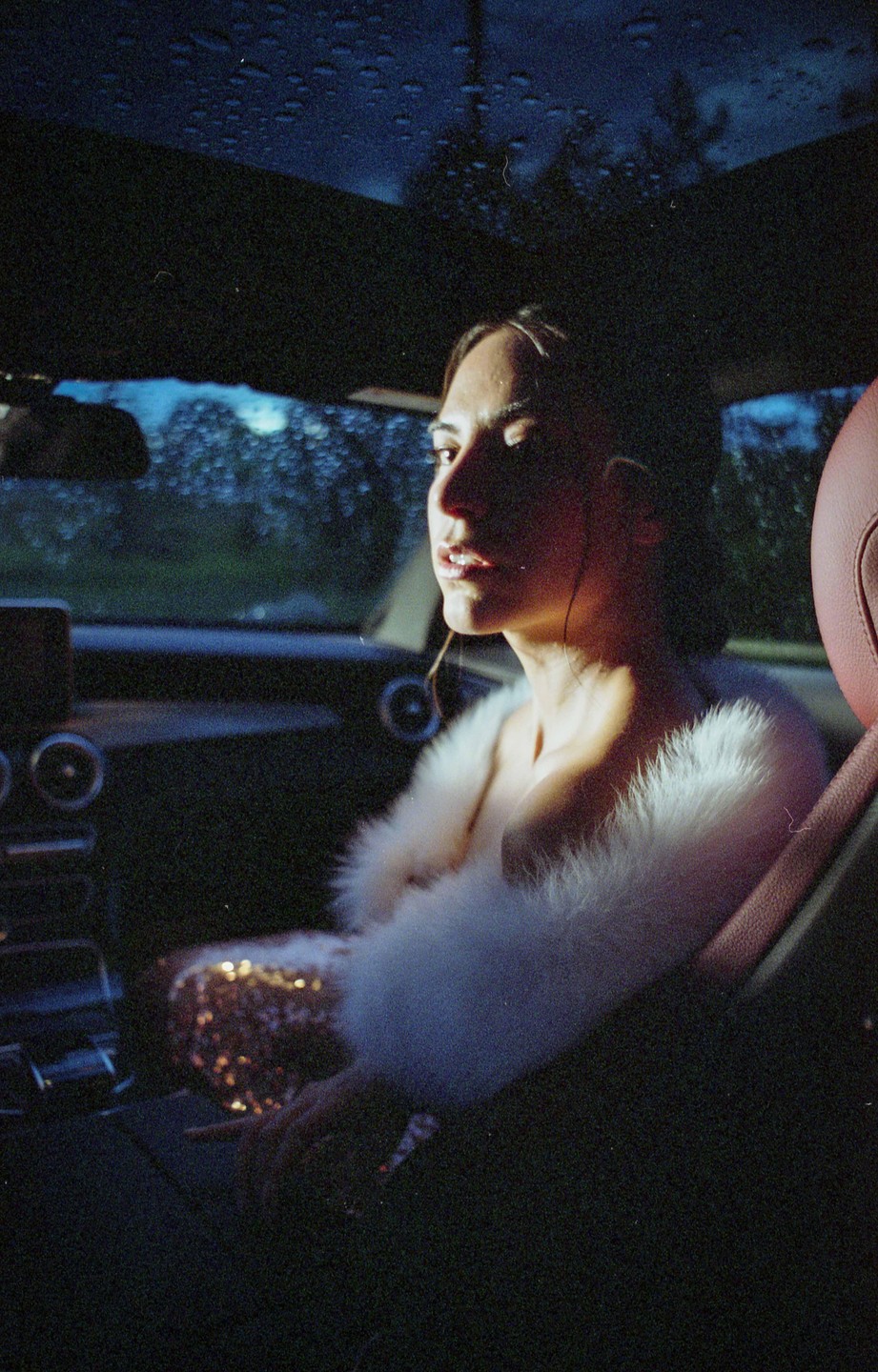 A woman in a white feathered top and a sequined dress sits in a car at night, with raindrops visible on the windshield.