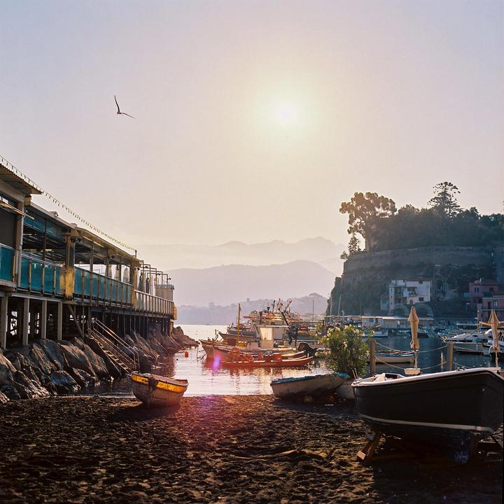 A bird flies over a beach with boats and a building on stilts, with mountains in the distance.