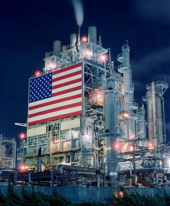 A large American flag is displayed on the side of an industrial refinery at night.