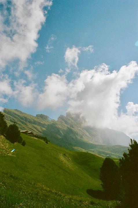 A green mountain slope with scattered trees and buildings is visible under a blue sky with white clouds.