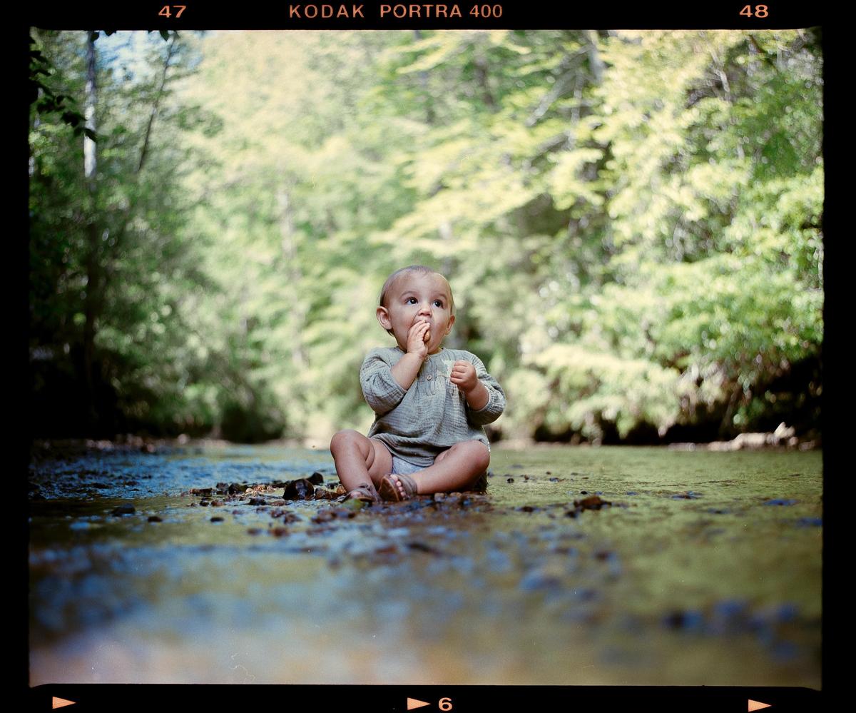 A baby sits in a shallow creek with trees in the background.