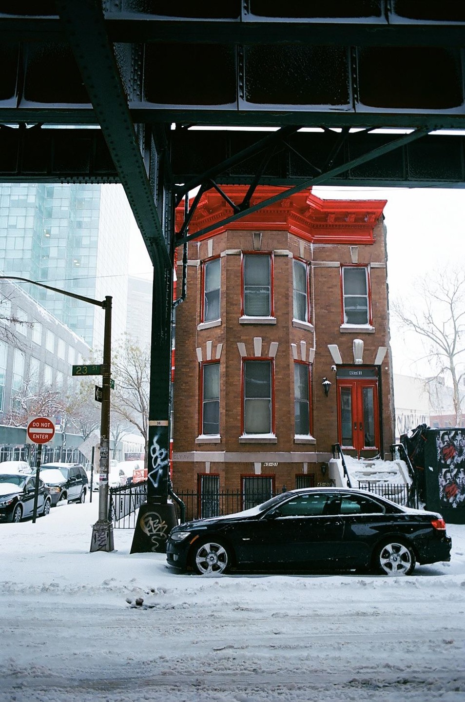 A black car is parked on a snow-covered street next to a brick building with red trim, under an elevated train track.