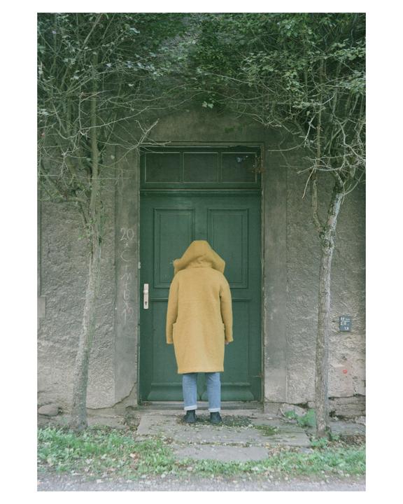 A person in a yellow coat stands facing a green door with trees on either side.