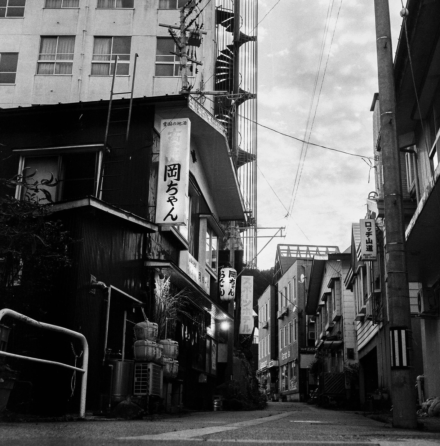 A narrow street in Minami Uonuma City is lined with buildings and utility poles under a cloudy sky.