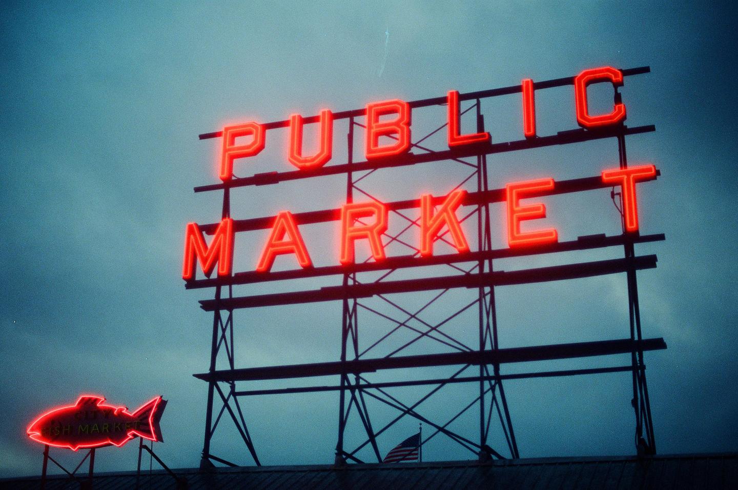A neon sign for a public market and a fish market stands against a cloudy sky.
