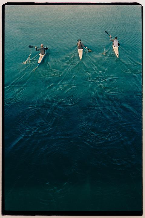 Three people in kayaks paddle across a body of water, viewed from above.