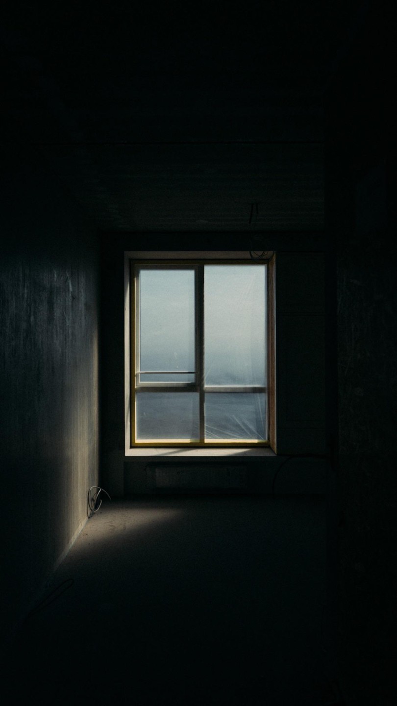Sunlight streams through a window covered with plastic sheeting into a dark, empty room.