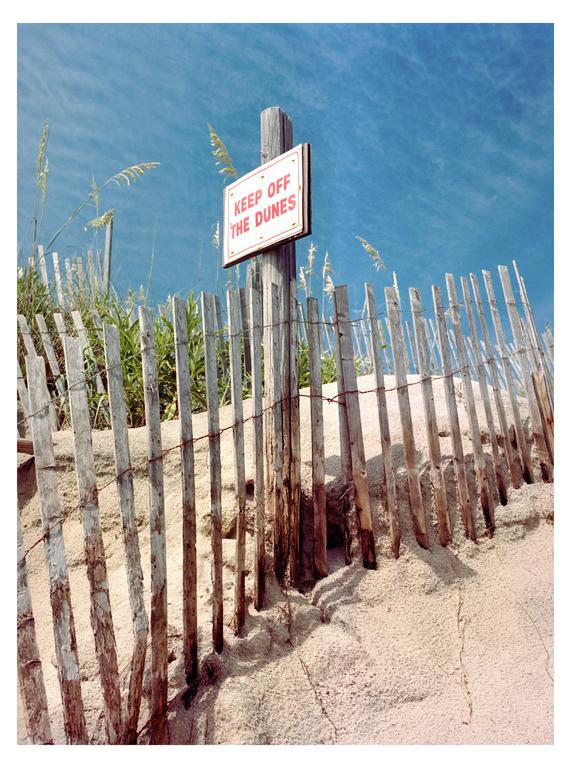 A wooden fence with a "Keep Off The Dunes" sign stands on a sandy dune under a blue sky.