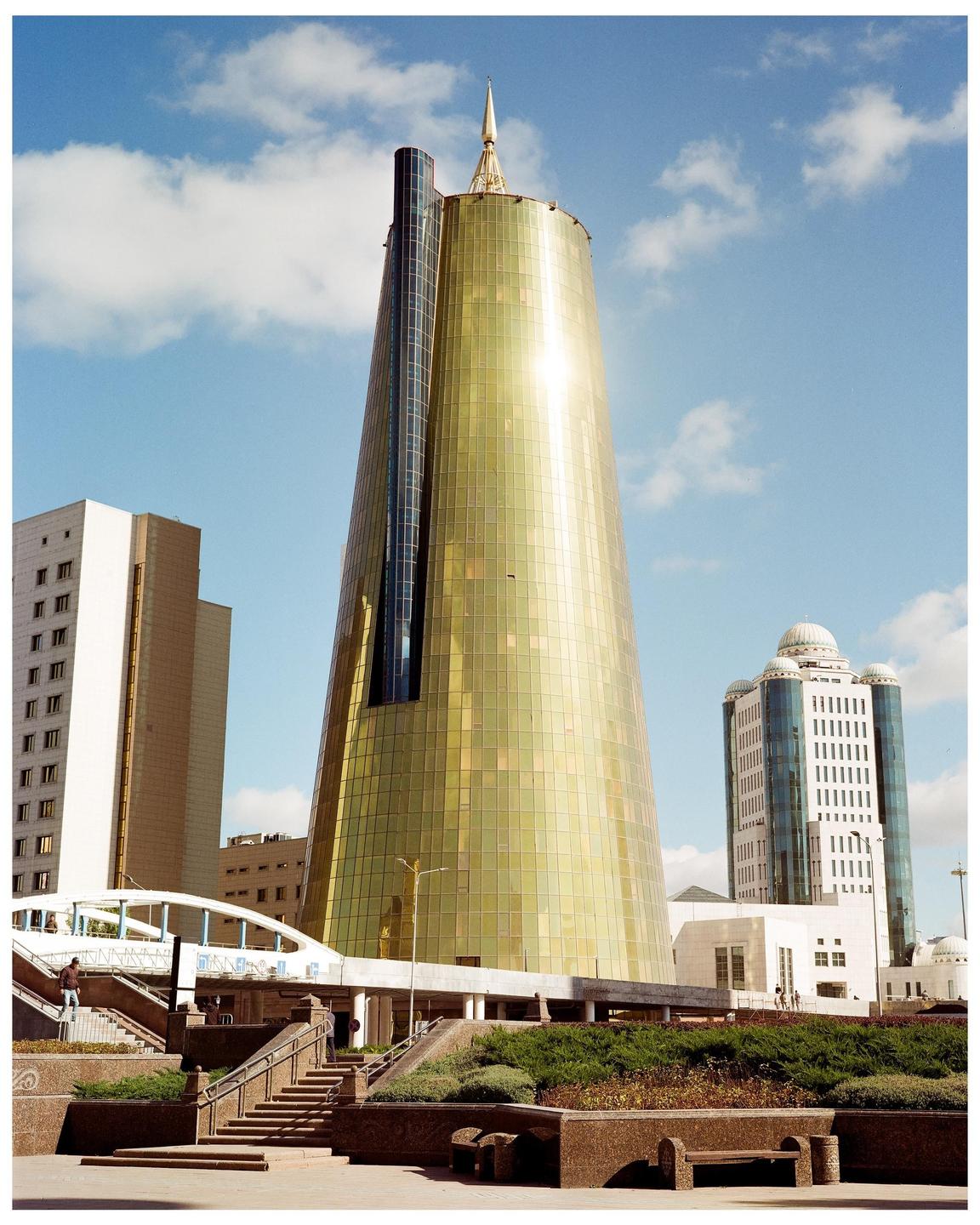 A tall, golden, conical building with a spire stands prominently against a blue sky with scattered clouds, surrounded by other buildings and a pedestrian bridge.
