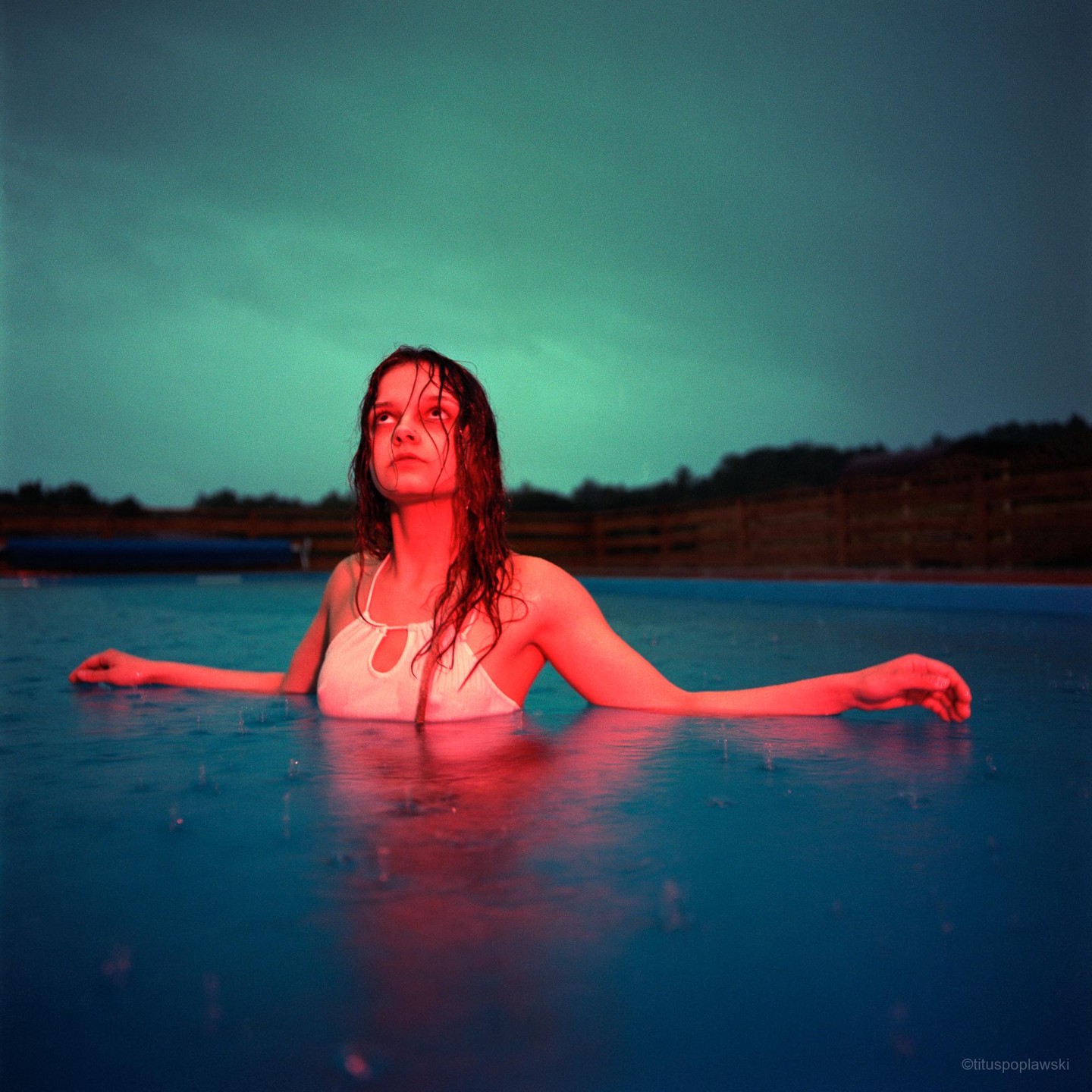 A woman with wet hair is in a swimming pool with red light on her body and a green sky above.