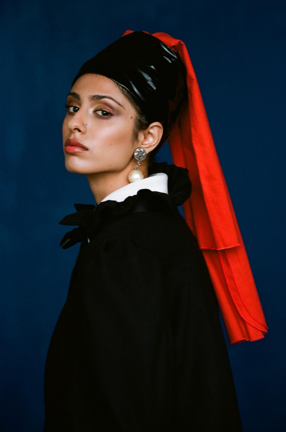 A woman wearing a black head covering with a red scarf and a black cape with a white collar and a pearl earring looks at the viewer.
