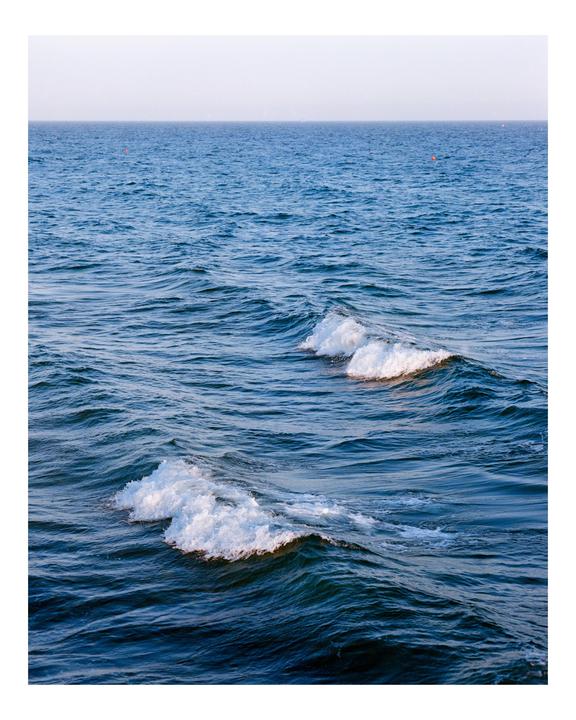 Two small waves with white foam crests move across a dark blue ocean under a pale sky.