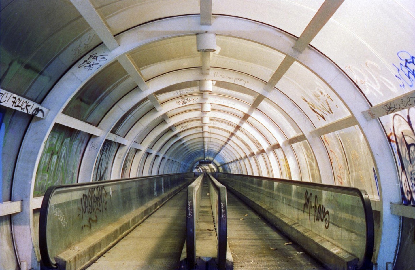 A long, empty, graffiti-covered moving walkway extends into the distance within a tunnel.