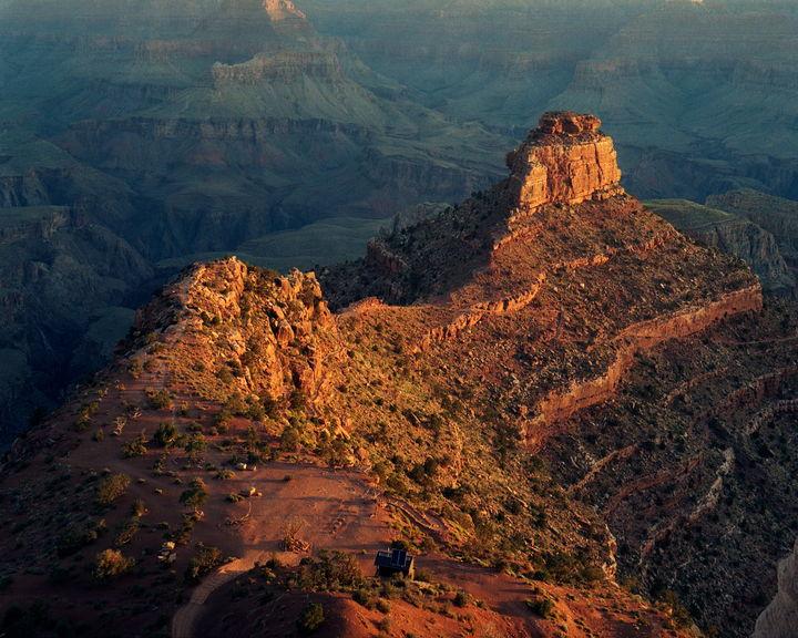 A small building sits on a sunlit ridge overlooking a vast canyon.