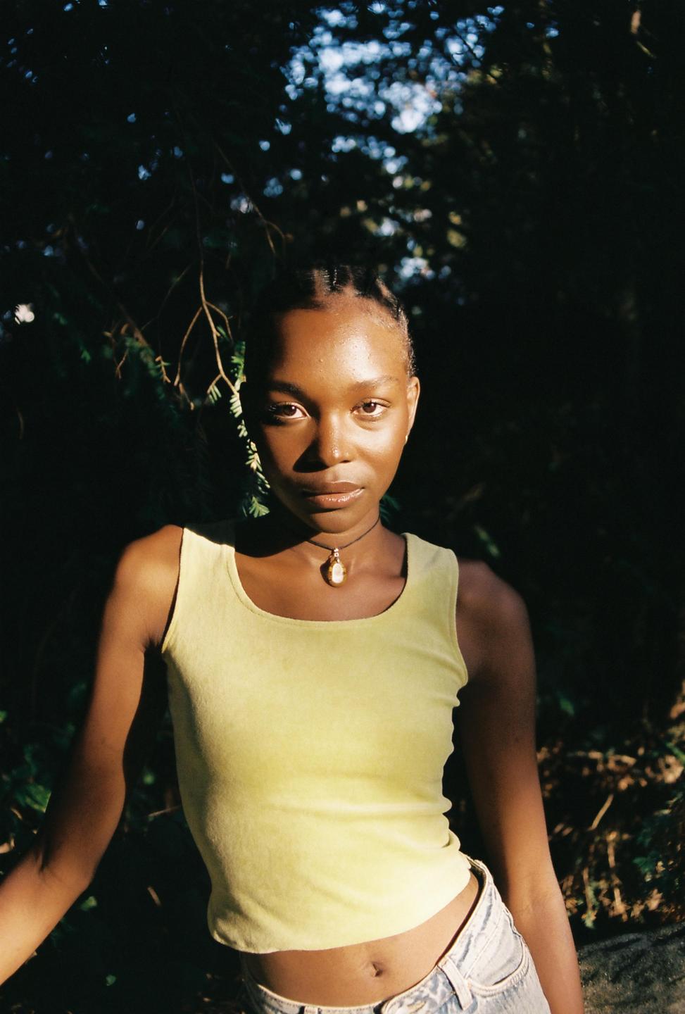A woman wearing a yellow top and a necklace is looking at the viewer in a sunlit outdoor setting.