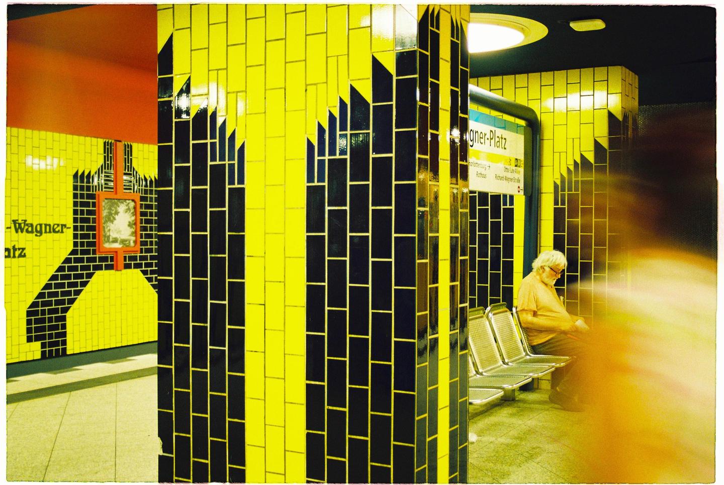A man sits on a bench in a subway station with yellow and black tiled walls.