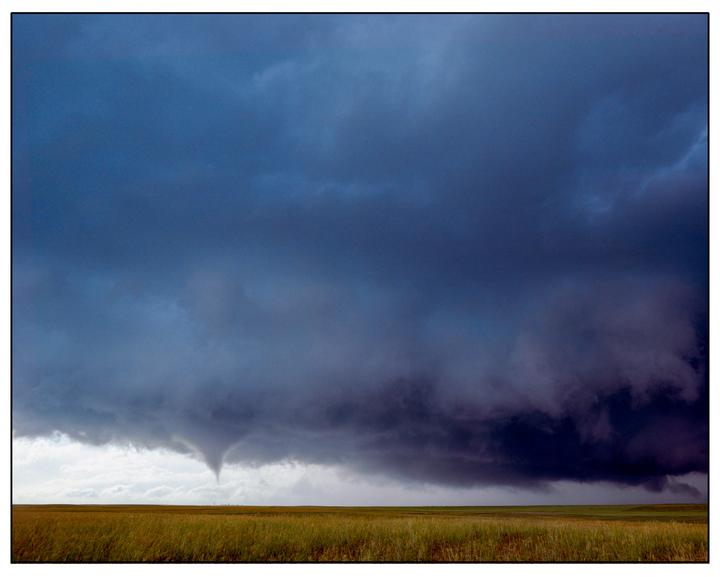 A tornado forms under a dark, stormy sky above a wide, grassy field.