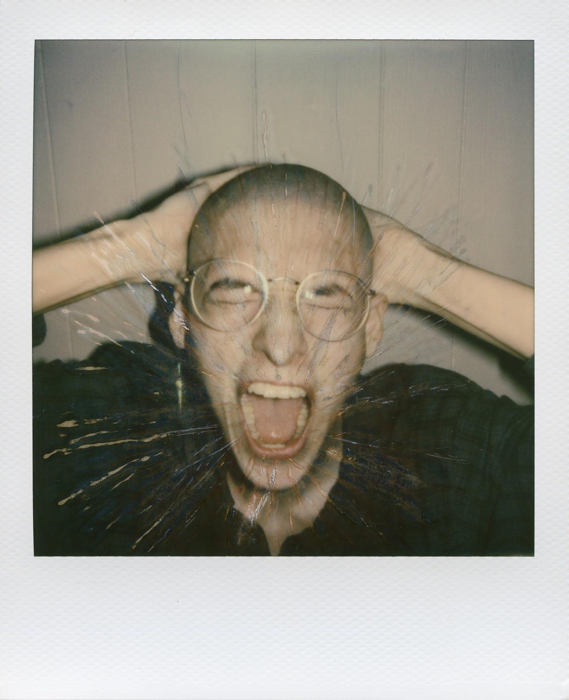 A person with a shaved head and glasses screams with hands on their head, covered in a splattered effect.