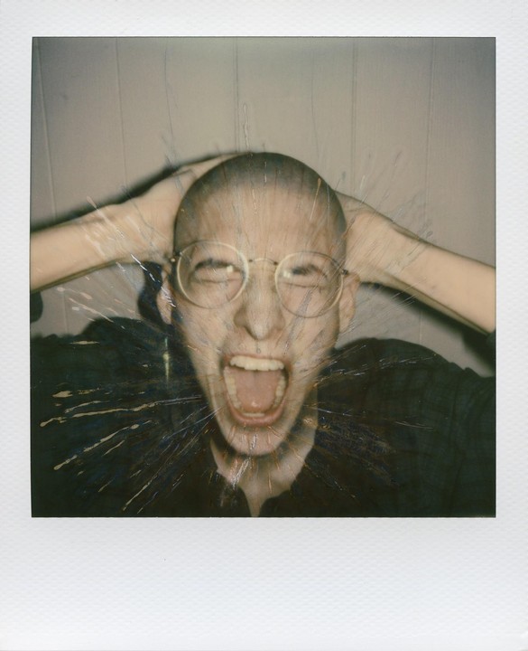 A person with a shaved head and glasses screams with hands on their head, covered in a splattered effect.