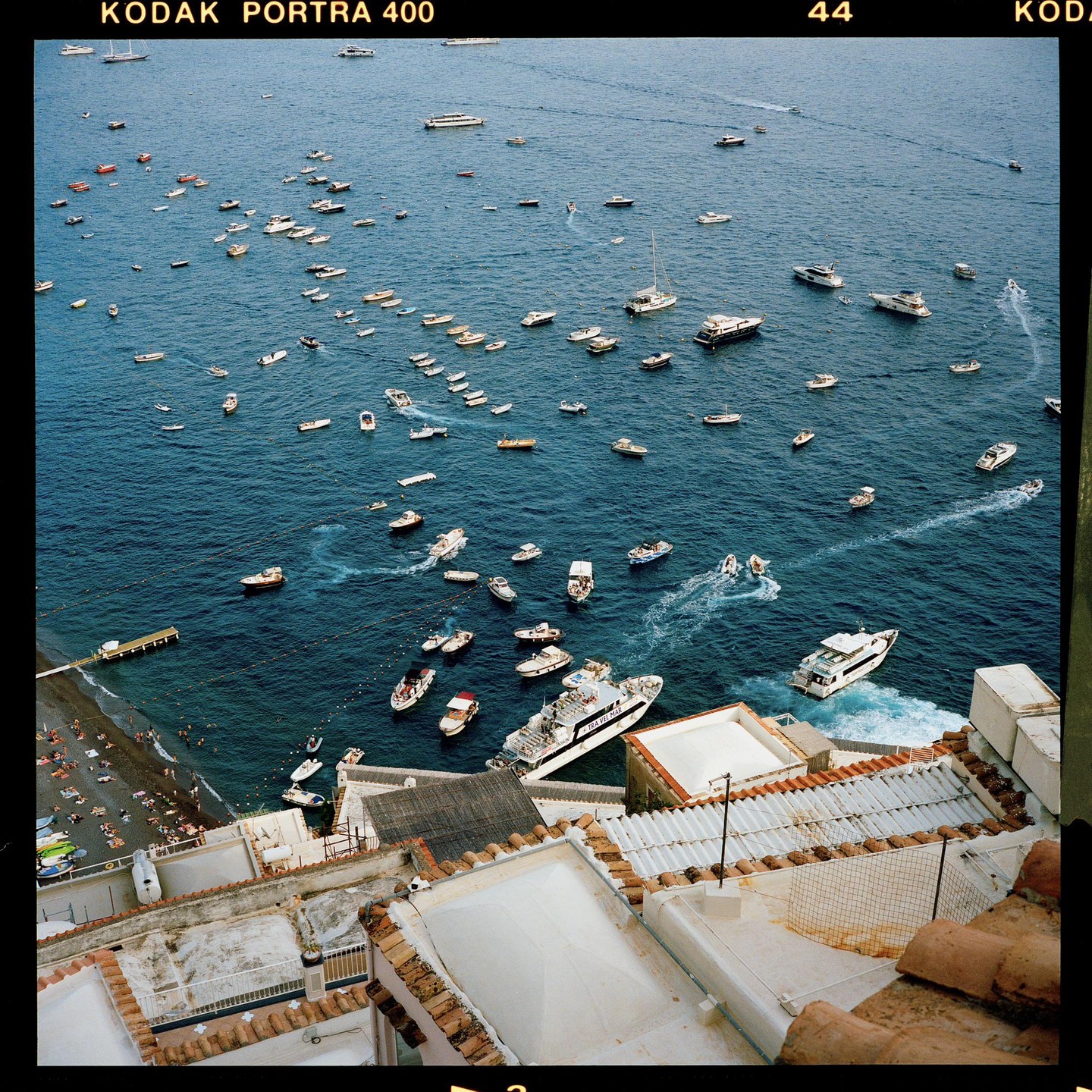 A high-angle view shows many boats and yachts on the blue sea next to a beach with sunbathers and buildings with terracotta roofs.