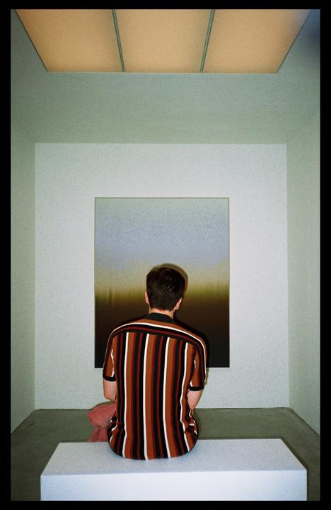 A man sits on a bench facing a painting in a museum.