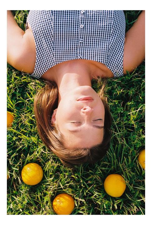 A woman with closed eyes lies in green grass surrounded by oranges.
