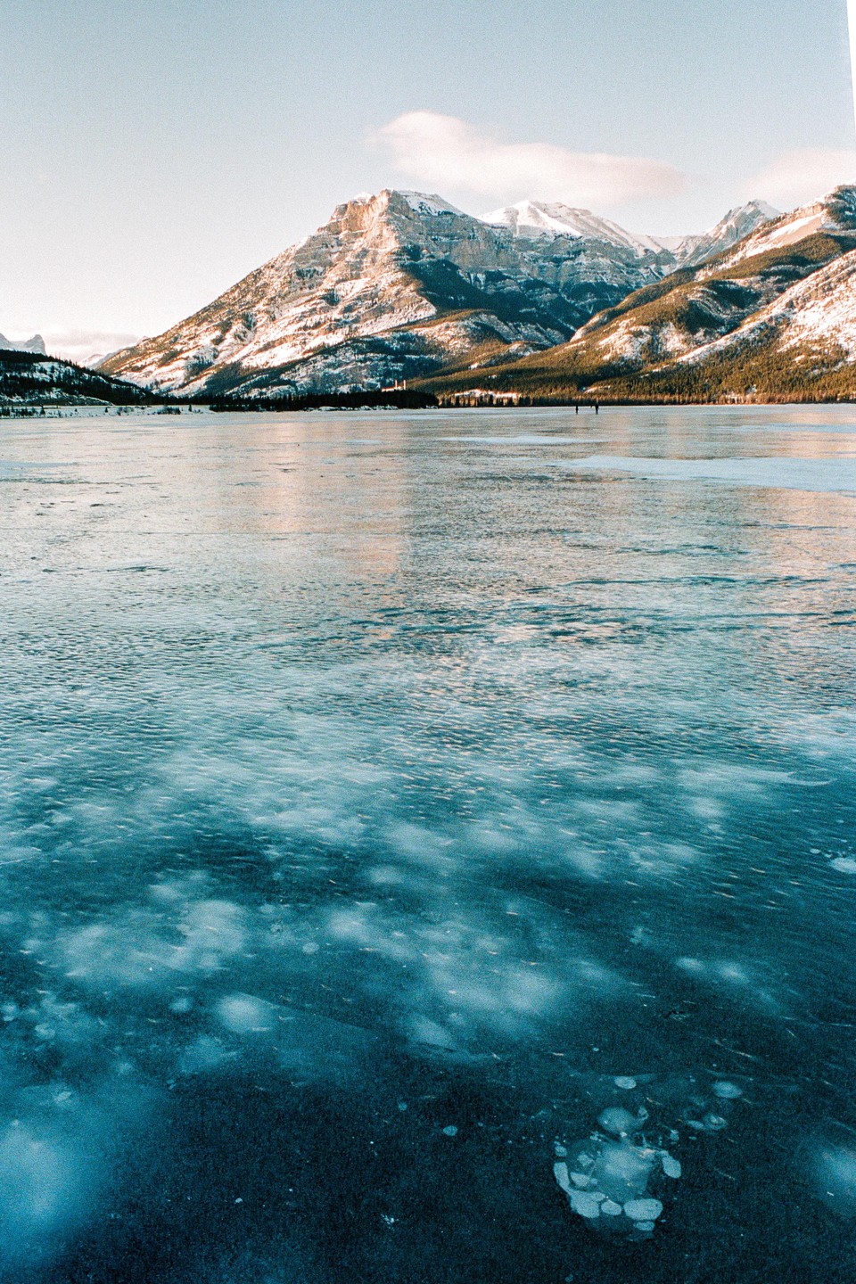 A frozen lake with methane bubbles is in the foreground, and snow-capped mountains are in the background.