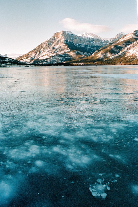 A frozen lake with methane bubbles is in the foreground, and snow-capped mountains are in the background.
