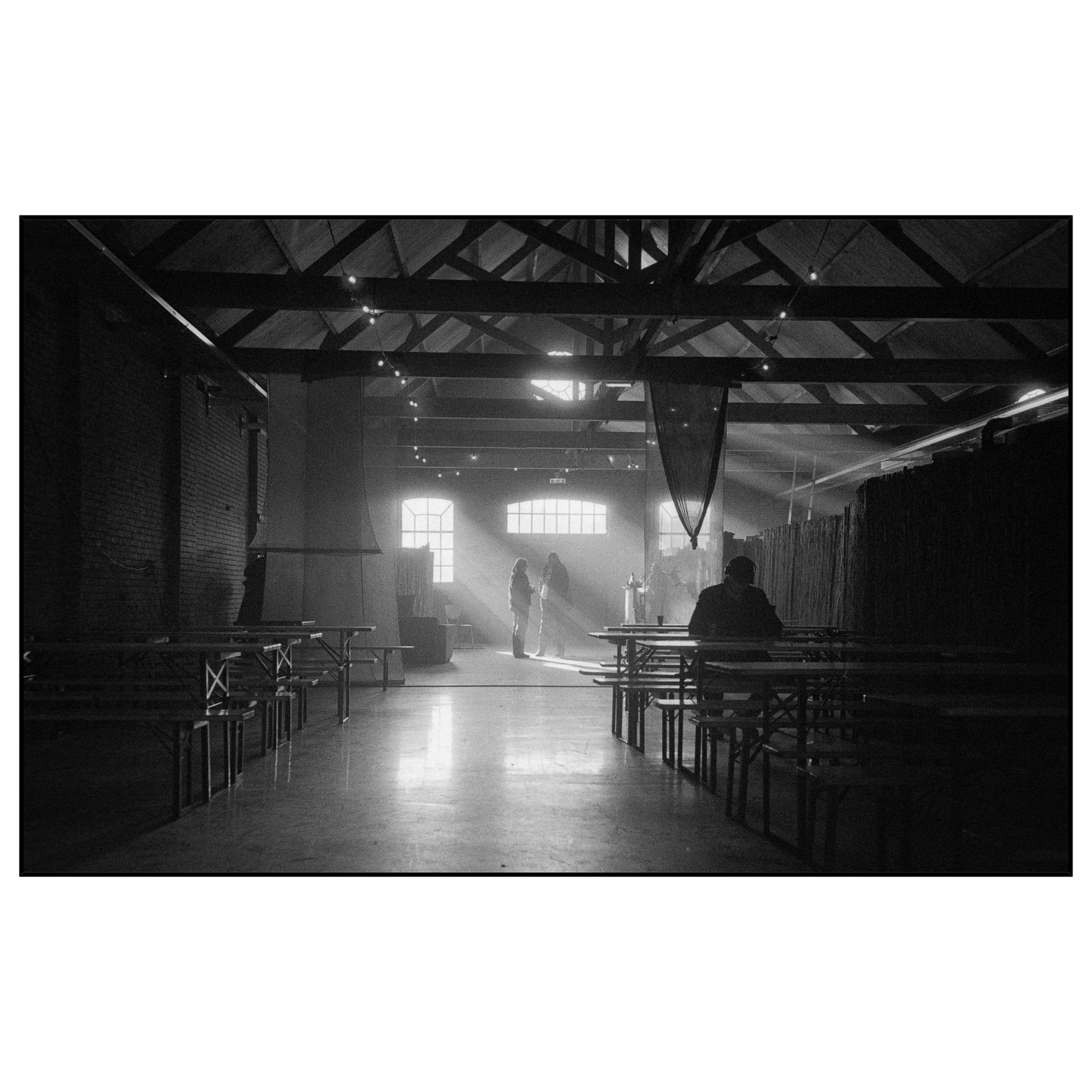 Sunlight streams through windows into a large, empty hall with tables and benches, illuminating two people standing in the distance and one man seated in the foreground.