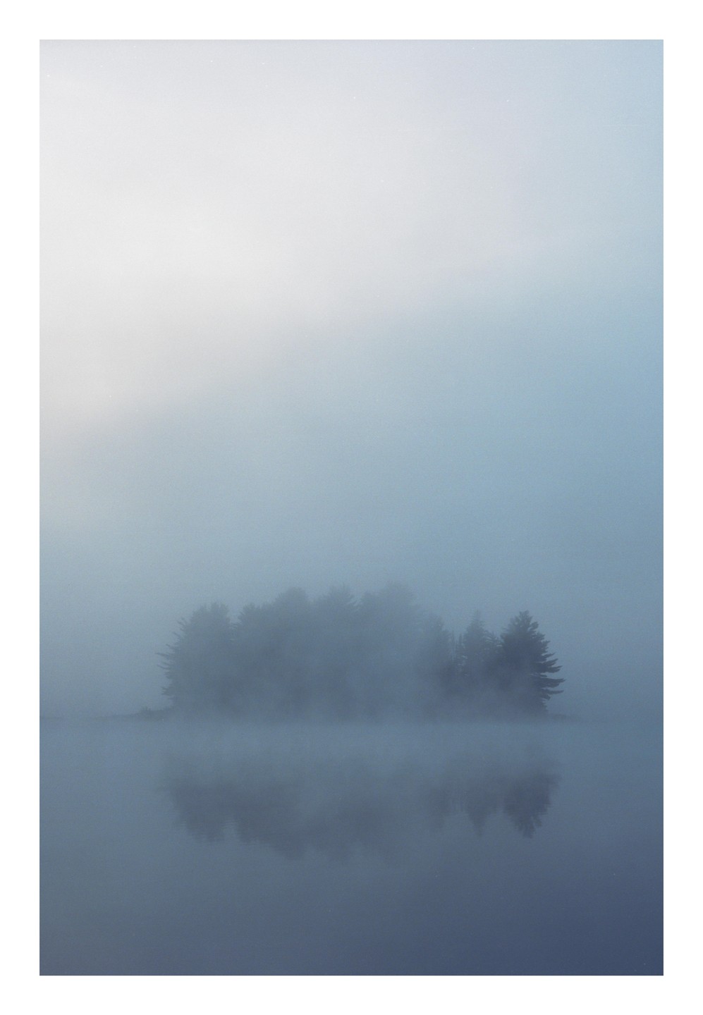 A small island of trees is shrouded in thick fog, reflecting faintly on the still water below.