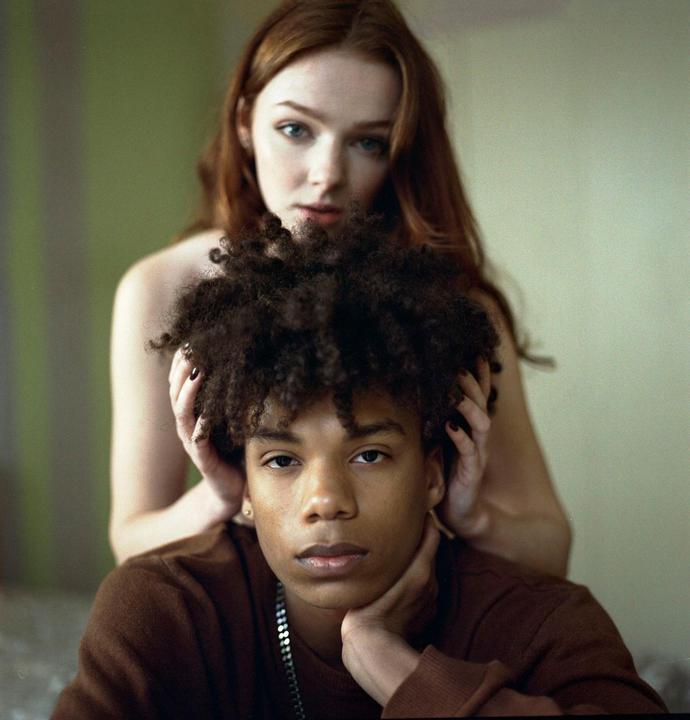 A woman with long red hair stands behind a man with curly hair who is resting his chin on his hand.