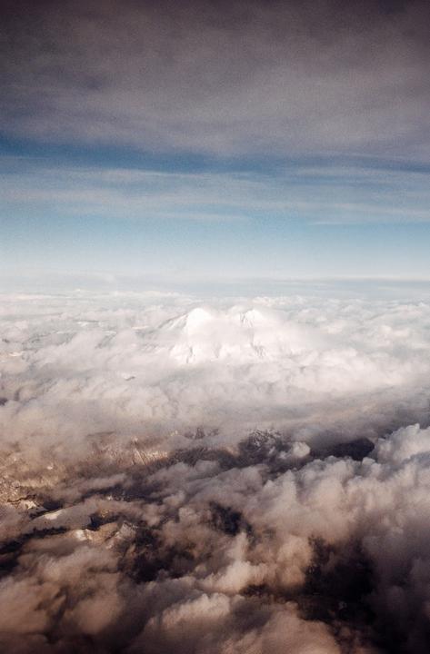 A snow-capped mountain peak rises above a sea of clouds, with a clear blue sky above.