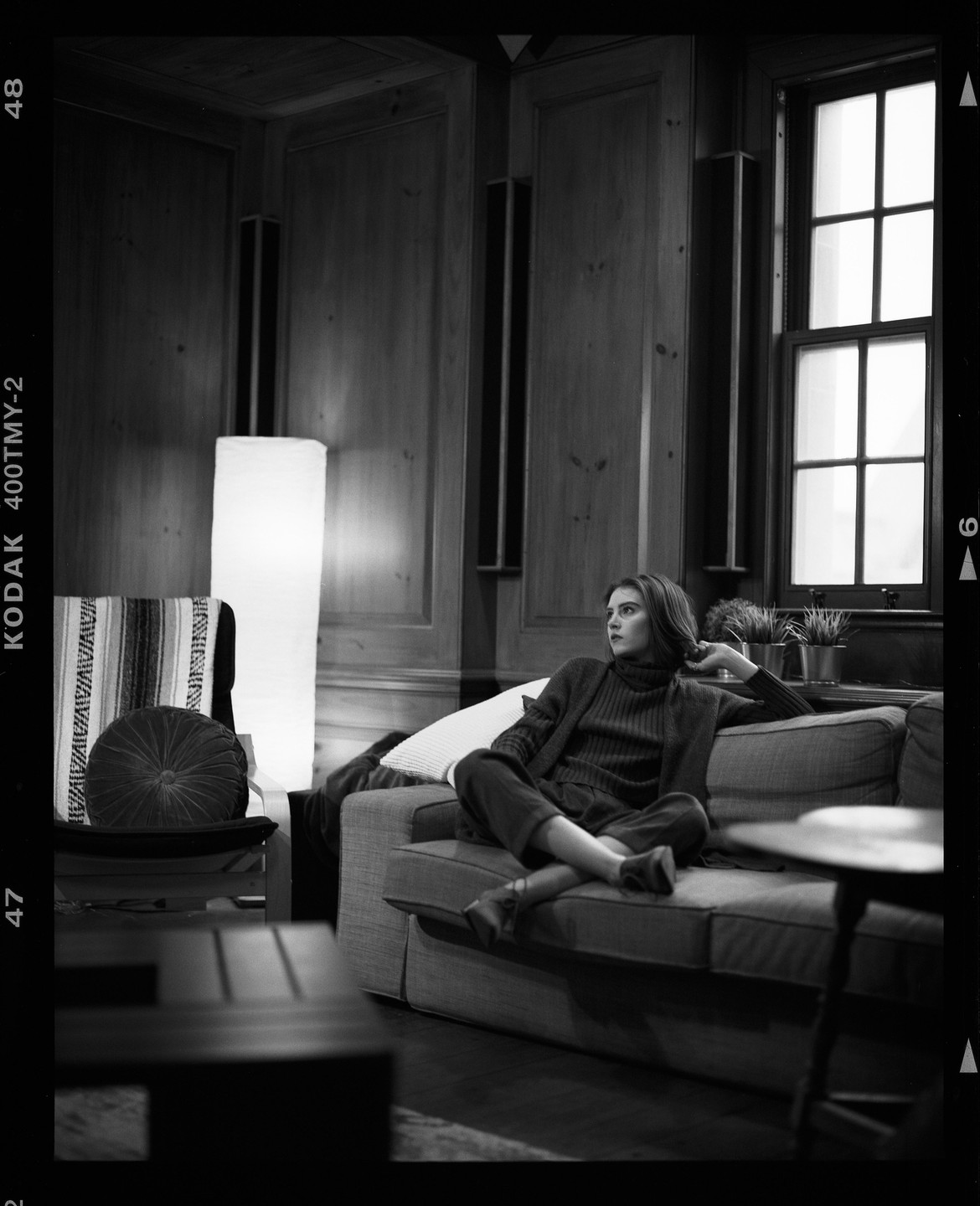 A woman sits on a sofa in a room with wood paneling and a large window.