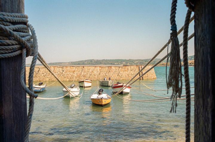 Small boats are moored in a harbor with a stone wall and a distant green hill under a clear sky.