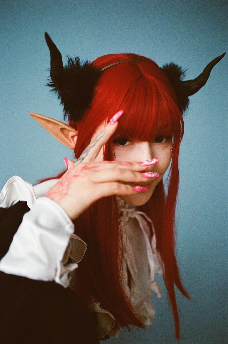 A woman with red hair, elf ears, and horns covers part of her face with her hand.