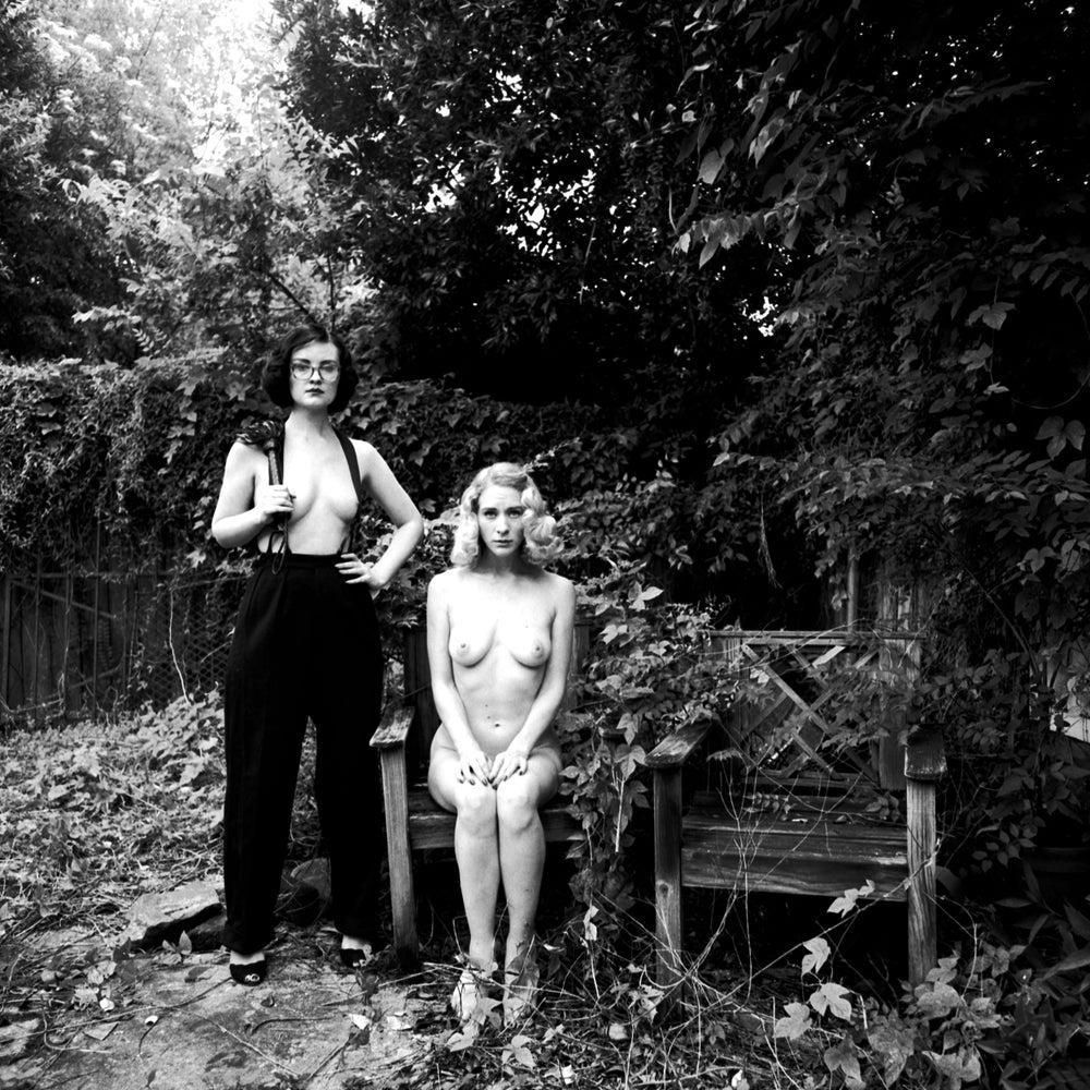 Two women pose outdoors, one standing in suspenders and trousers, the other sitting nude on a wooden bench.