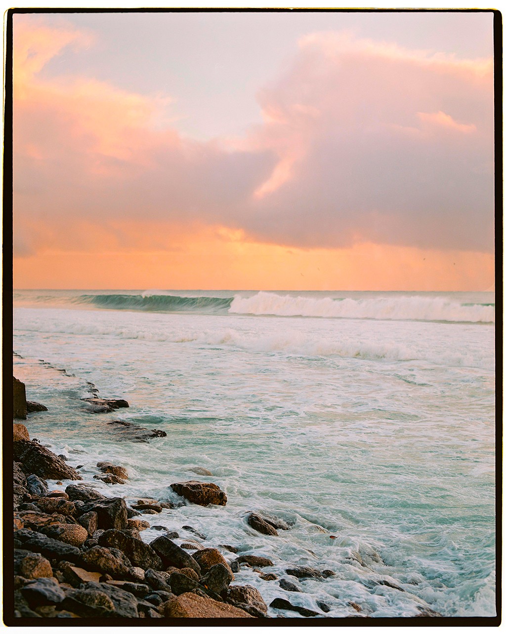 Waves crash on a rocky shore under a sky filled with pink and orange clouds.