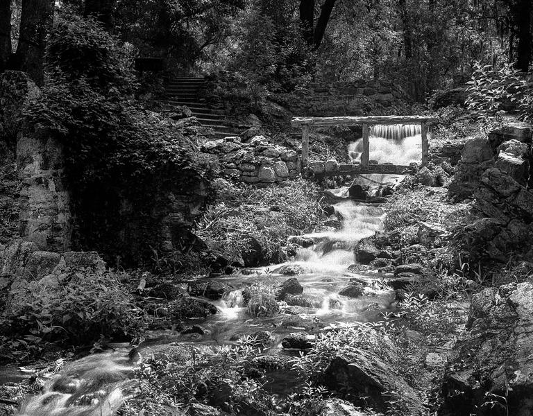 A small waterfall flows under a wooden bridge and down a rocky stream bed through a forest.